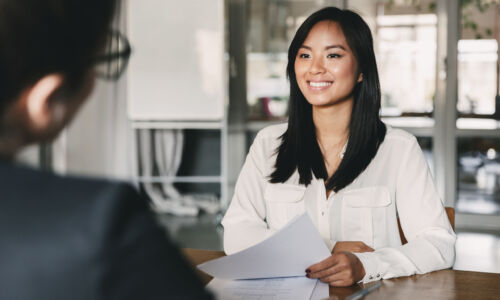 Portrait of joyful asian woman smiling and holding resume while sitting in front of businesswoman during corporate meeting or job interview - business, career and placement concept