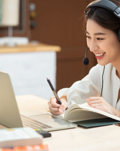 Smiling,Asian,Young,Female,Using,Headset,Looking,At,Laptop,Screen