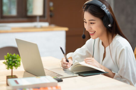 Smiling,Asian,Young,Female,Using,Headset,Looking,At,Laptop,Screen